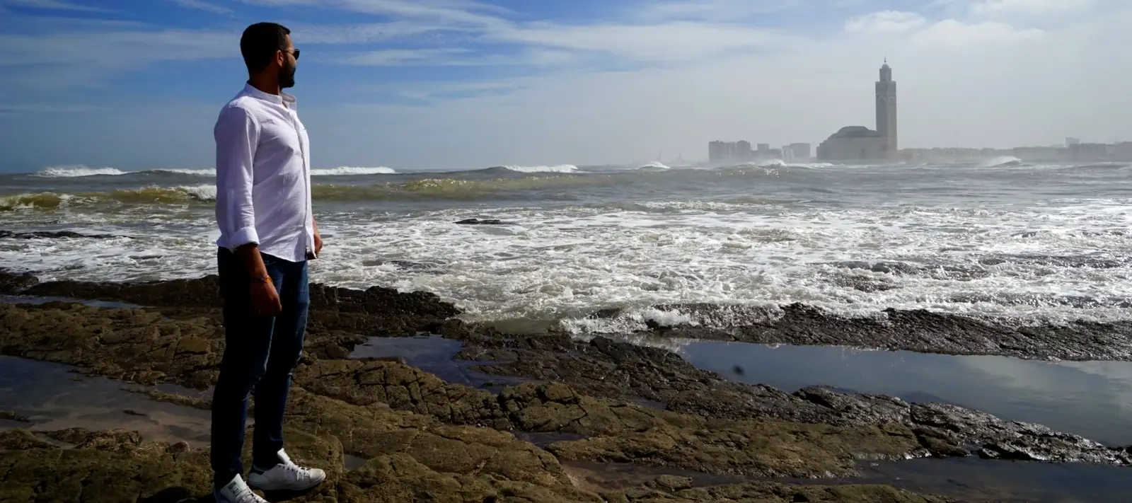 Reda on the rocks at the Casablanca shoreline, with the Hassan II Mosque minaret across the water.